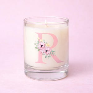 May include: A clear glass candle holder with a white candle inside. The candle is decorated with a pink letter "R" and a floral design. The candle sits on a pink surface.