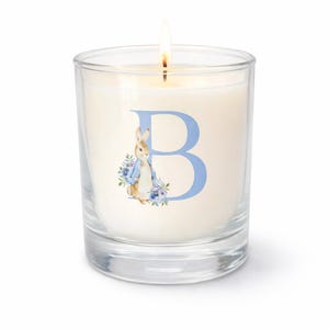 May include: A lit candle in a clear glass votive. The candle has a cream-colored wax and features a blue letter "B" with a watercolor illustration of a rabbit and flowers. The candle is on a white background.