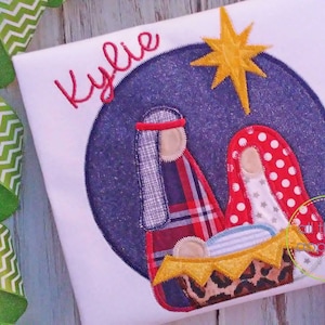 May include: A white t-shirt with a blue, red, and white applique of the nativity scene. The name "Kylie" is embroidered in red above the scene. A yellow star is above the scene.