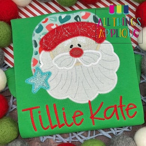 May include: Green t-shirt with an embroidered applique of a Santa Claus face with a blue star. The name "Tillie Kate" is embroidered in red below the applique.