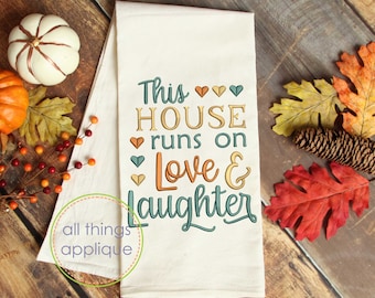 Könnte beinhalten: Ein weißes Küchentuch mit dem Text "This house runs on love & laughter" in grüner, orangefarbener und brauner Schrift mit kleinen Herzen zwischen den Wörtern. Das Tuch ist von Herbstdekorationen umgeben, darunter Kürbisse, Blätter und Tannenzapfen.