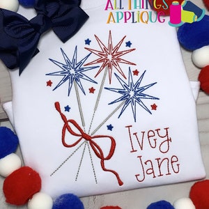 May include: White t-shirt with red, white, and blue embroidered fireworks and a red bow. The name "Ivey Jane" is embroidered below the fireworks. The text "All Things Applique" is in the top right corner of the image.