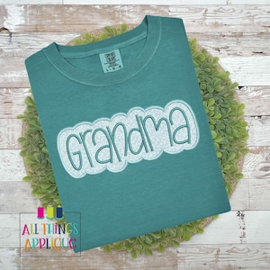 Puede incluir: Una camiseta azul turquesa con la palabra "Grandma" en una fuente bordada blanca y azul.