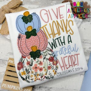 May include: A white shirt featuring a design of three pumpkins stacked on top of each other. The pumpkins are decorated with floral patterns and polka dots. The text "Give thanks with a grateful heart" is written in a cursive font above the pumpkins. The text "Colossians 3:15-16" is written below the pumpkins.