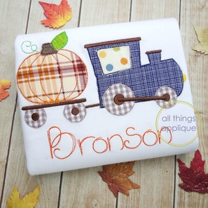 May include: A white onesie with an embroidered pumpkin train design. The train is blue with white and brown wheels. The pumpkin is orange and brown with a green stem. The onesie has the name "Bronson" embroidered in orange.