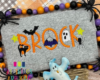 May include: A gray t-shirt with the name "Brock" embroidered in orange with Halloween-themed embellishments including a black cat, bats, a ghost, and a spider.