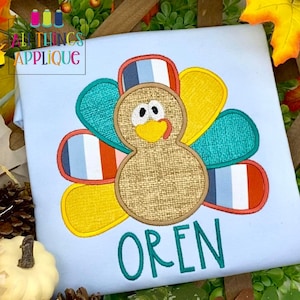 May include: A light blue t-shirt with an embroidered turkey design. The turkey has a brown body, yellow beak, and a red tongue. The turkey's feathers are multi-colored and striped. The name "OREN" is embroidered in green below the turkey.