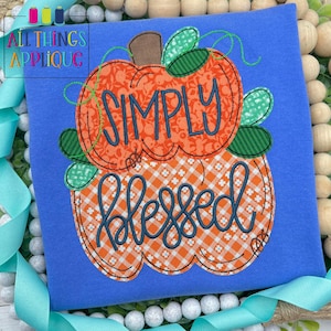 May include: A blue t-shirt with an appliqued pumpkin design. The top pumpkin has the word "SIMPLY" in blue, and the bottom pumpkin has the word "BLESSED" in blue. The pumpkins are orange with green leaves and brown stems.