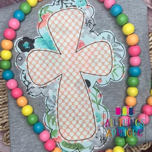 May include: A light gray t-shirt with a peach cross applique with white polka dots. The cross is surrounded by a blue and white floral design. The text "All Things Applique" is visible in the bottom right corner of the image.