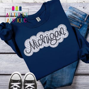 以下が含まれることがあります： 白い星で囲まれた「ミシガン」という文字がグレーと白で刺繍された、ネイビーブルーのスウェットシャツです