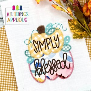 Puede incluir: Toalla de cocina de lino blanco con un diseño de calabaza. La calabaza se divide en dos secciones, una con un patrón de cuadros amarillos y blancos y la otra con un patrón floral. Las palabras "SIMPLY blessed" están bordadas en la calabaza.