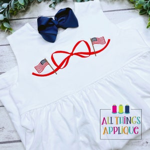 K&ouml;nnte beinhalten: Wei&szlig;es &auml;rmelloses Kleid mit einer roten Schleife und zwei gestickten amerikanischen Flaggen auf der Vorderseite. Das Kleid hat einen R&uuml;schen-Saum. Der Text "All Things Applique" ist in der rechten unteren Ecke sichtbar.