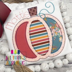 May include: A white shirt with a pink and red pumpkin applique. The pumpkin has a brown and blue striped pattern, a floral pattern, and a brown stem with white polka dots. The shirt also has the name "Mattie" embroidered on it. The shirt is from All Things Applique.
