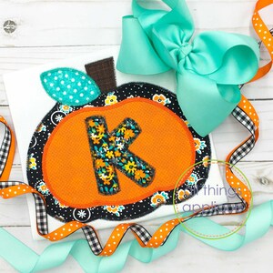 May include: A white shirt with a black and orange pumpkin applique featuring the letter K. The pumpkin is decorated with floral fabric and has a green leaf. The shirt is surrounded by teal, orange, and black and white ribbon.