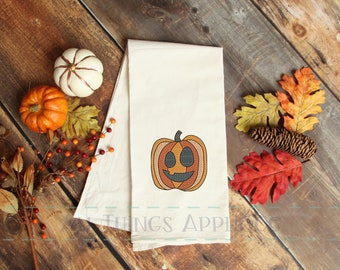 Puede incluir: Una toalla de cocina blanca con una calabaza bordada de color naranja, amarillo y negro con una cara sonriente. La toalla está sobre una superficie de madera con decoraciones de otoño.