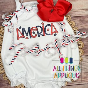 May include: White baby onesie with red, white, and blue ribbon. The onesie has the word "America" embroidered on it in red and blue with white stars. The onesie is on a wooden surface with a red bow.