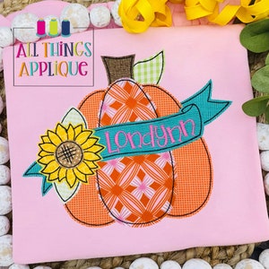 May include: A pink t-shirt with an embroidered applique of a pumpkin with a sunflower and a banner that says "Londyn".
