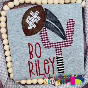 May include: A gray shirt with a red and white gingham football goal post and a brown football with white stitching. The name "Bo Riley" is embroidered in red on the shirt.