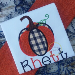 Puede incluir: Una camiseta blanca con una aplicación de calabaza a cuadros en blanco y negro y el nombre "Rhett" en letras rojas y negras.
