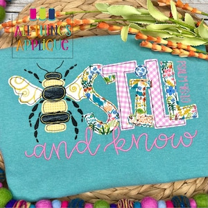 May include: A turquoise t-shirt with an embroidered bee and the words "STILL and know" in pink script. The letters of "STILL" are filled with floral patterns and pink gingham.