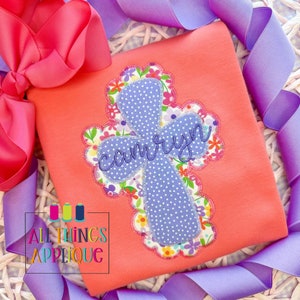 Könnte beinhalten: Ein rosa Shirt mit einem lilafarbenen Kreuz-Applikationen mit weißen Punkten und einem floralen Rand. Das Kreuz ist mit dem Namen "carolyn" bestickt.