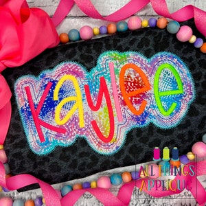 May include: A colorful, rainbow-colored, embroidered name "Kaylee" on a black fabric with a leopard print. The name is outlined in a bright blue, green, and orange thread. The fabric is surrounded by pink and white ribbon and colorful beads.