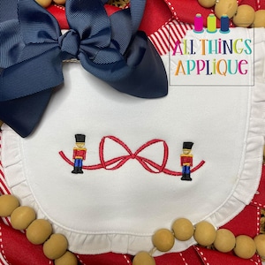 May include: White bib with a red bow and two toy soldier figures embroidered on it. The bib has a ruffled edge. The text "ALL THINGS APPLIQUE" is visible in the top right corner of the image.
