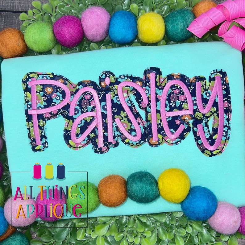 May include: A turquoise shirt with the name "Paisley" embroidered in pink and navy blue floral fabric. The name is in a large, stylized font. The shirt is surrounded by colorful pom-poms.