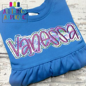 May include: A blue sweatshirt with a floral patterned applique of the name "Vanessa" in a pink outline.