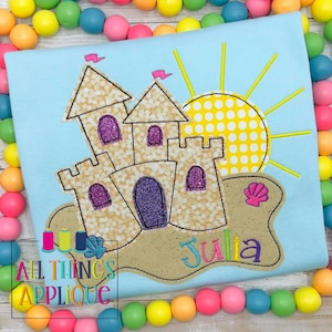 May include: A light blue t-shirt with an embroidered design of a sandcastle, sun, and seashell. The name "Julia" is embroidered below the sandcastle. The t-shirt is surrounded by a colorful beaded garland.