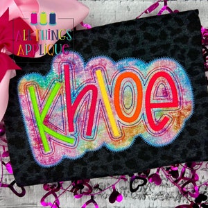 May include: A black shirt with a rainbow tie-dye design featuring the name "Khloe" in a colorful, multi-layered font.