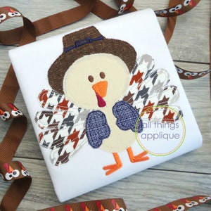 Könnte beinhalten: Eine braune und weiße Truthahn-Applikation mit einem braunen Hut und blauen Flügeln. Der Truthahn trägt eine blaue Fliege. Die Applikation befindet sich auf einem weißen Shirt. Der Text "all things applique" befindet sich in einem Kreis auf dem Shirt.