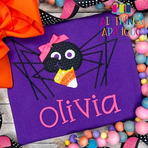 May include: Purple shirt with a black spider holding a candy corn. The spider has a pink bow on its head and the shirt says "Olivia" in pink letters.