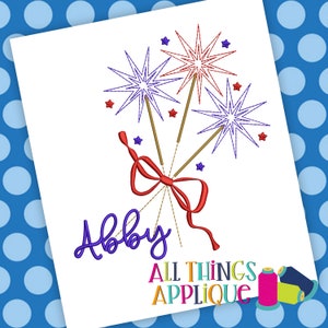 May include: Embroidered design of three sparklers tied together with a red bow. The name "Abby" is embroidered below the sparklers. The text "All Things Applique" is at the bottom of the design.