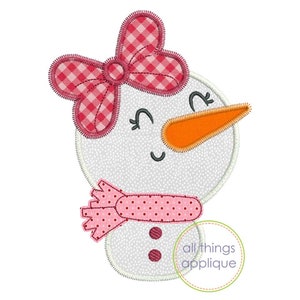 Pode incluir: Uma aplicação de boneco de neve branco com um laço xadrez rosa e branco, uma echarpe de bolinhas rosa e um nariz de cenoura. O boneco de neve tem dois botões pretos e um rosto sorridente.