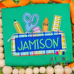 May include: Green t-shirt with a blue and white checkered pencil case with the name Jamison embroidered on it. The pencil case has a zipper and is filled with a pencil, scissors, a ruler, and a crayon.