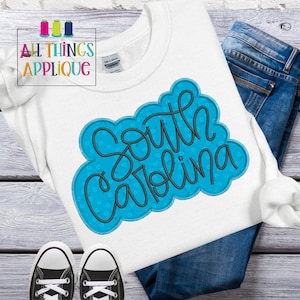 May include: White t-shirt with a blue and black appliqué that says "South Carolina" in a cursive font.