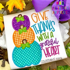 Puede incluir: Una camiseta blanca con una pila de tres calabazas con estampado de cuadros. Las calabazas son moradas, naranjas y azul verdoso. El texto "Give Thanks with a grateful heart" está escrito en una fuente de letra cursiva de colores. El texto "Colossians 3:15-16" está escrito en una fuente más pequeña debajo del texto principal.