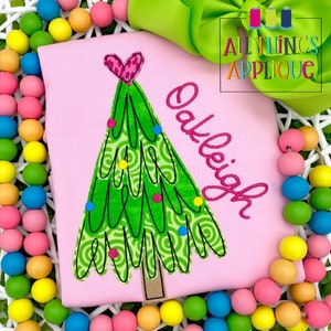 May include: Pink shirt with a green Christmas tree applique with a pink heart on top. The name "Oakleigh" is embroidered in pink on the shirt.