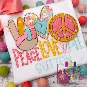 May include: A white shirt with an embroidered design featuring a peace sign, a heart, a pie, and the text "Peace Love & Pie Sutton". The design is in shades of pink, yellow, blue, and green. The shirt is from All Things Applique.
