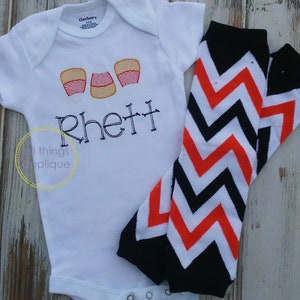 May include: White baby onesie with three orange and white candy corn appliques and the name "Rhett" embroidered in black. The onesie is paired with black and orange chevron leg warmers.