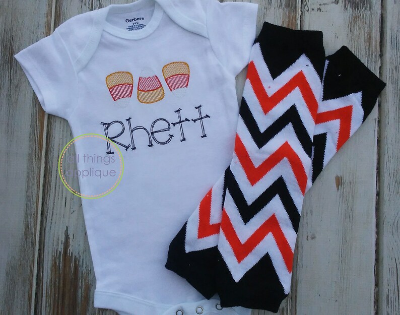 May include: White baby onesie with three orange and white candy corn appliques and the name "Rhett" embroidered in black. The onesie is paired with black and orange chevron leg warmers.
