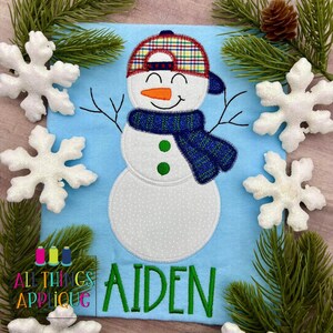 Op de afbeelding: Een lichtblauwe stof met een sneeuwman-applicatie. De sneeuwman heeft een geruite hoed en een blauwe sjaal, met een oranje wortelneus. De naam "Aiden" is eronder groen geborduurd. Decoratieve sneeuwvlokken en dennentakken omringen de applicatie.