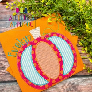 以下が含まれることがあります： オレンジ色のTシャツに、刺繍されたかぼちゃのデザイン。かぼちゃは、ピンクのアウトライン、青と白のストライプ、オレンジ色の水玉模様の異なる色の生地で作られています。シャツには緑色の刺繍で「エブリン」という名前が書かれています。