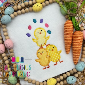 May include: A yellow chick juggling colorful Easter eggs. The chick is standing on top of two other chicks. The image is surrounded by a border of wooden beads and Easter eggs.