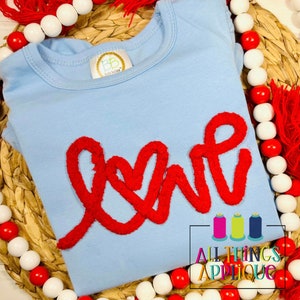 May include: A light blue t-shirt with a red fuzzy applique of the word "love" in a cursive font. The shirt is surrounded by red and white beads.