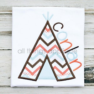 May include: A white tee shirt with an embroidered design of a brown and orange teepee with a blue and white chevron pattern. The name "Conlan" is embroidered in orange and blue on the right side of the teepee.