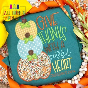 May include: Teal-colored t-shirt with an embroidered design of three stacked pumpkins. The pumpkins are decorated with various patterns and colors. The shirt features the text "GIVE THANKS WITH A GRATEFUL HEART" in orange and green.