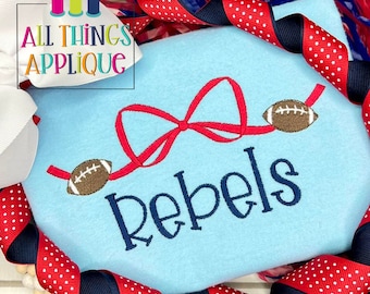 May include: A light blue t-shirt with a red bow and two brown footballs. The word "Rebels" is embroidered in navy blue below the bow. The t-shirt is surrounded by red and blue ribbon.