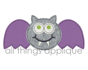 Puede incluir: Un murciélago de fieltro gris con alas de fieltro morado y una cara sonriente con dientes blancos. El murciélago tiene ojos verdes y una boca cosida.